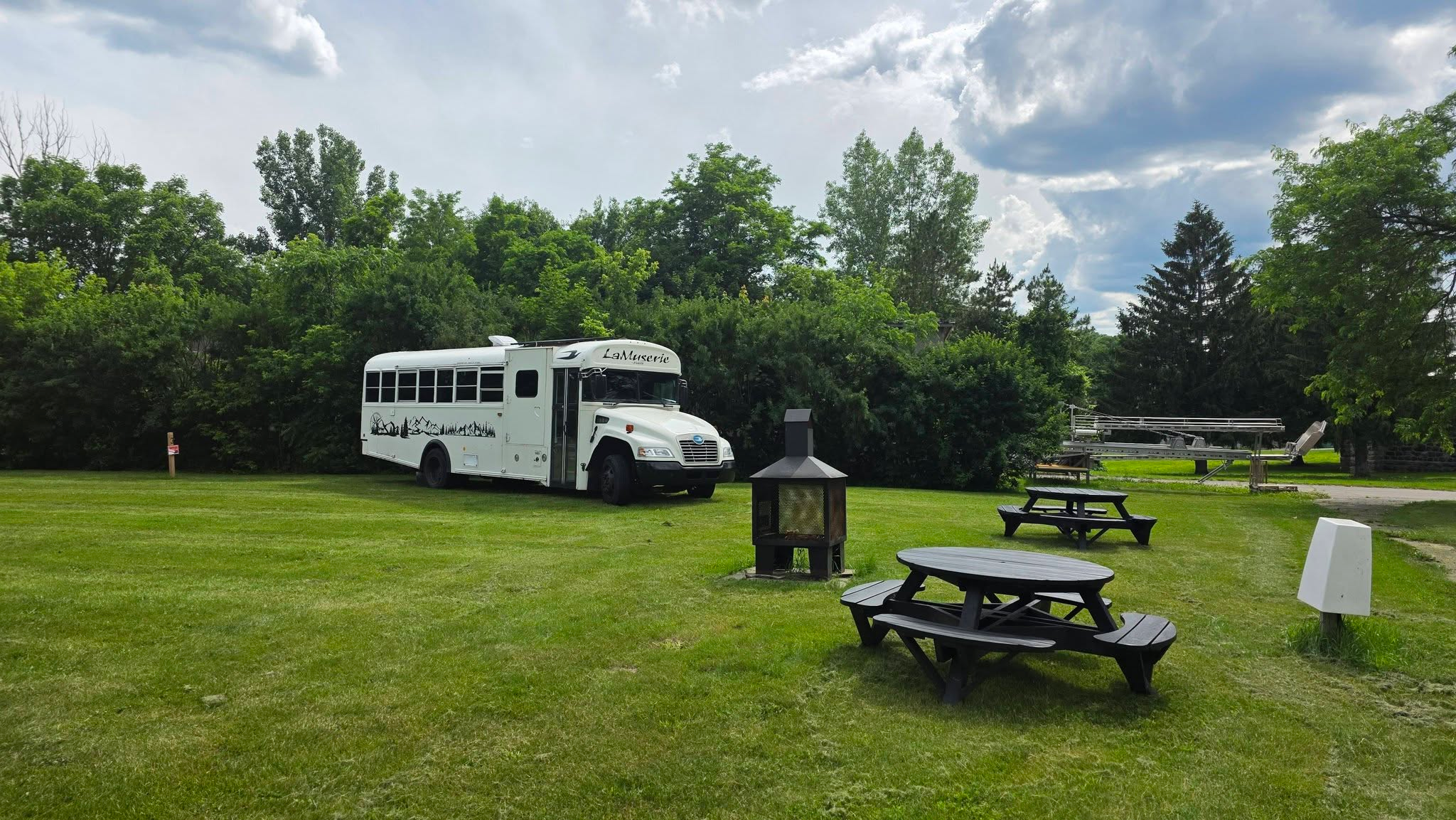 Terrain gazonné aménagé de l'Abbaye d'Oka avec un skoolie stationné près d'une espace feu et tables à picnics entourré d'arbres